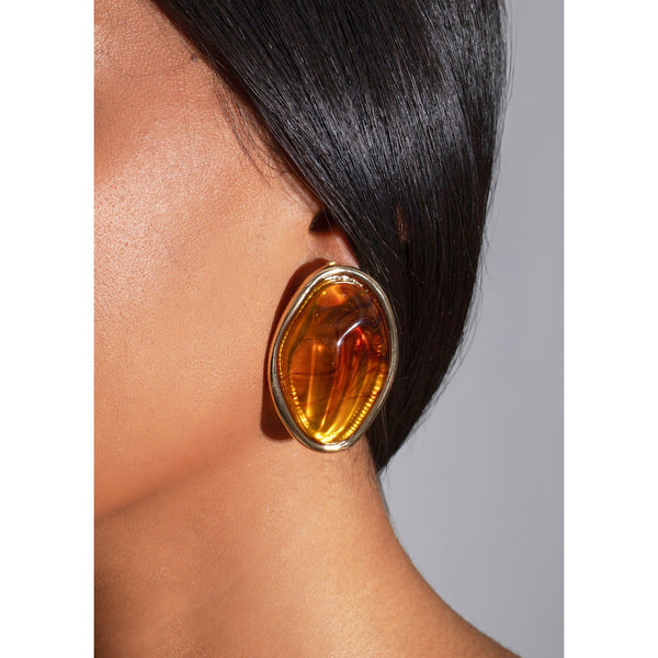 Close-up of a person wearing a gold earring with a amber-colored stone on a neutral background